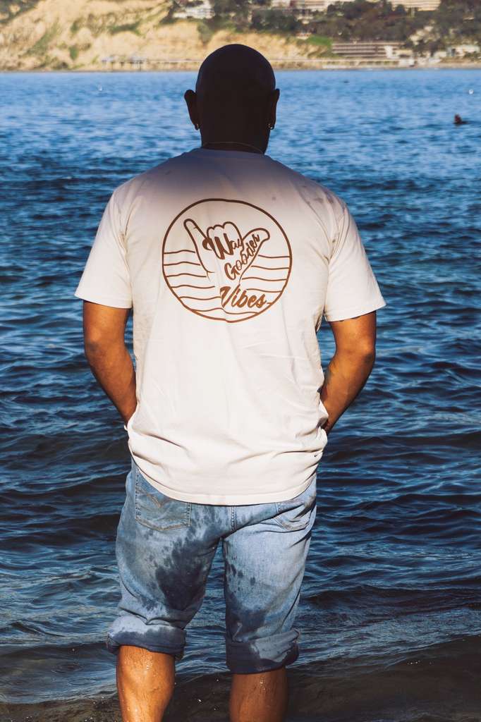 Man wearing the bone Gooder Vibes Tee near the coast, back view