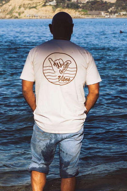 Man wearing the bone Gooder Vibes Tee near the coast, back view
